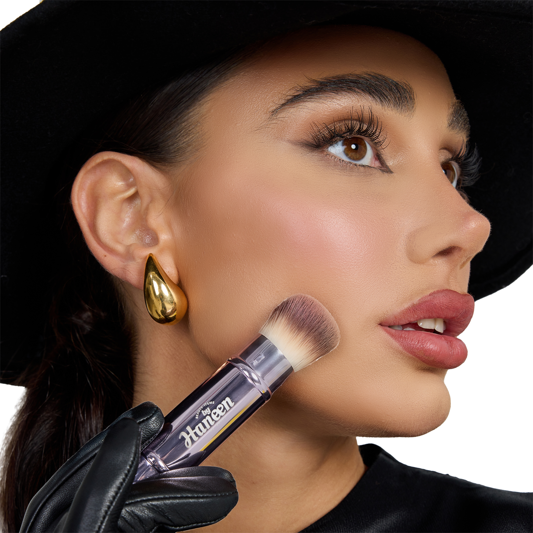 Woman holding a makeup brush with a visible brand name, wearing a black hat and gold earring.