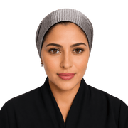 A woman wearing a gray ribbed knit head covering and a black garment, shown from the chest up against a solid black background.