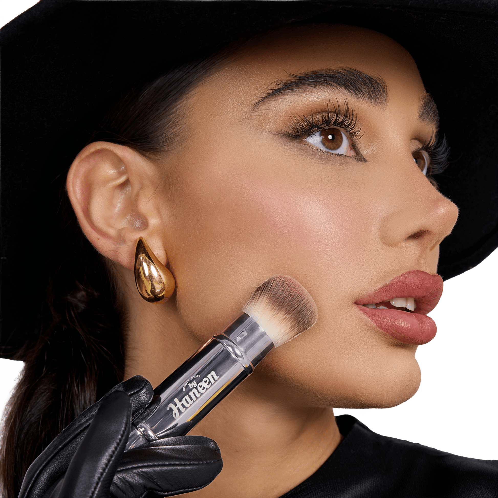 Woman holding a makeup brush with a visible brand name, wearing a black hat and gold earring.