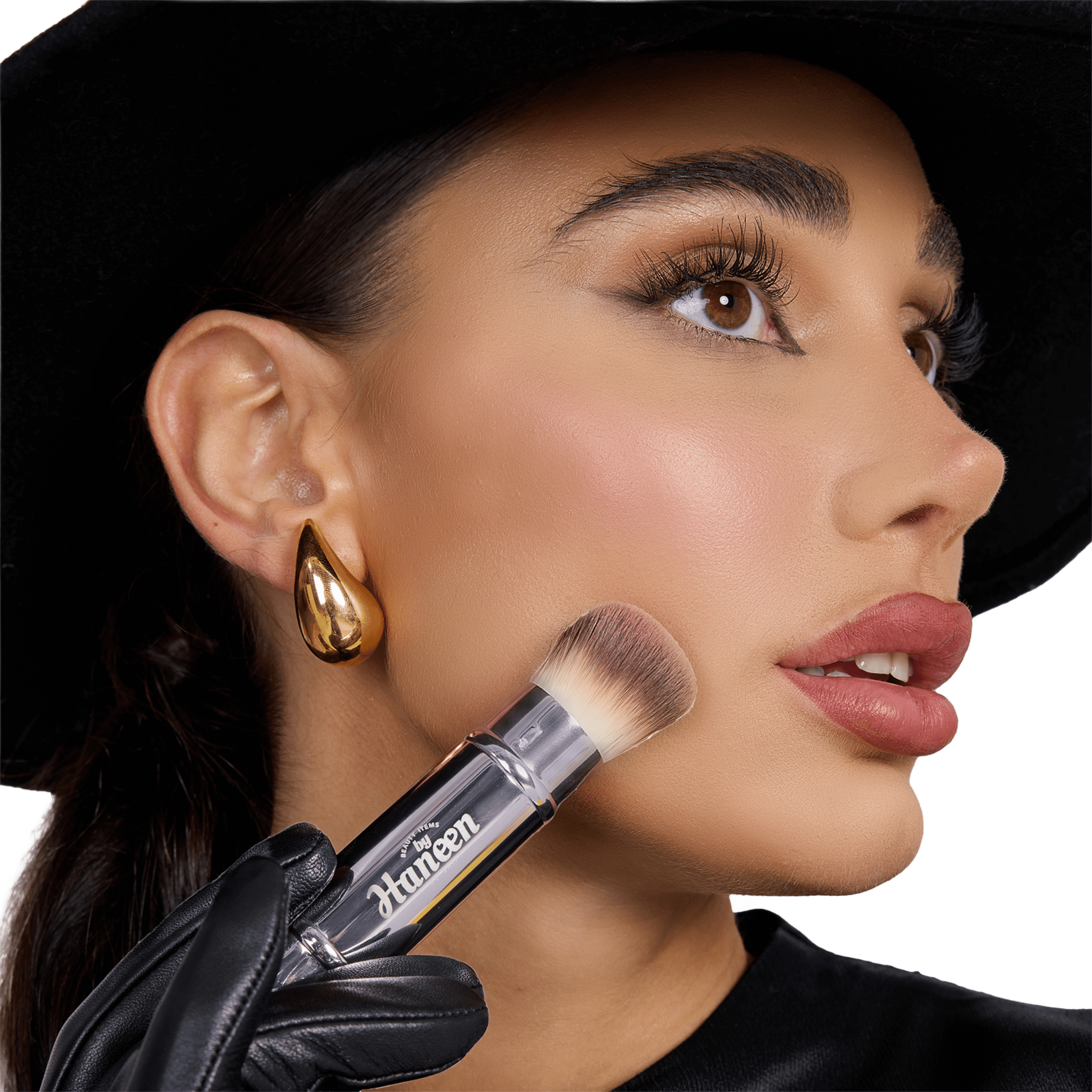 Woman holding a makeup brush with a visible brand name, wearing a black hat and gold earring.